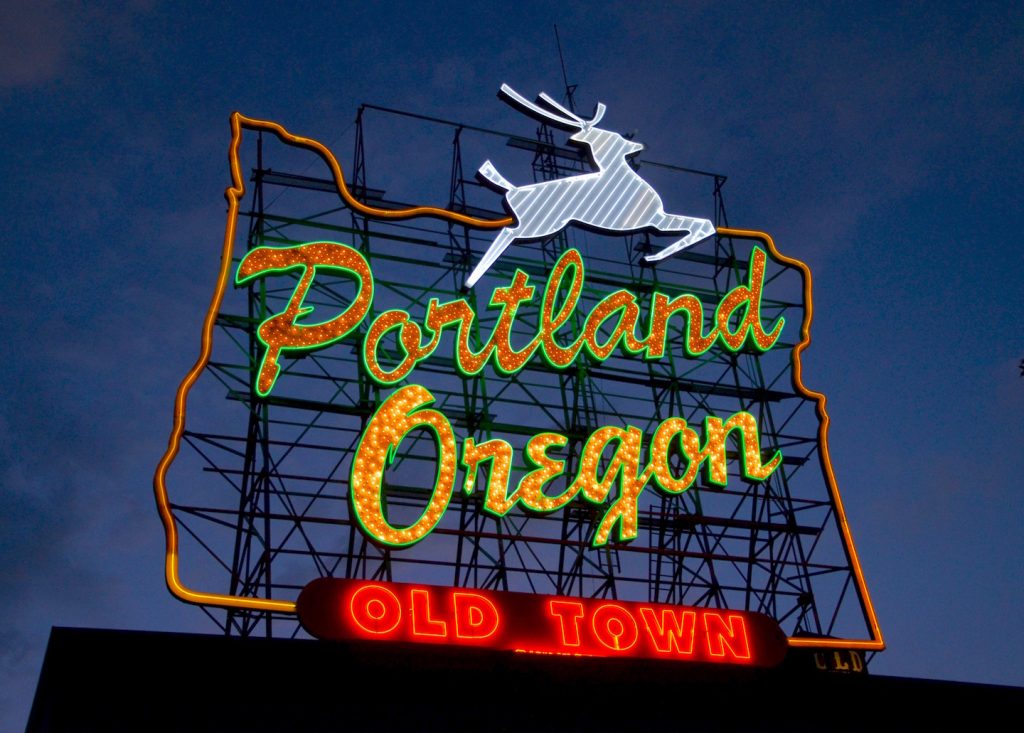 Photo of neon sign featuring the words "Portland Oregon" in green-outlined yellow-orange script and a white stag. The sign is outlined in the shape of Oregon. In smaller, all caps red text at the bottom of the sign are the words "Old Town"